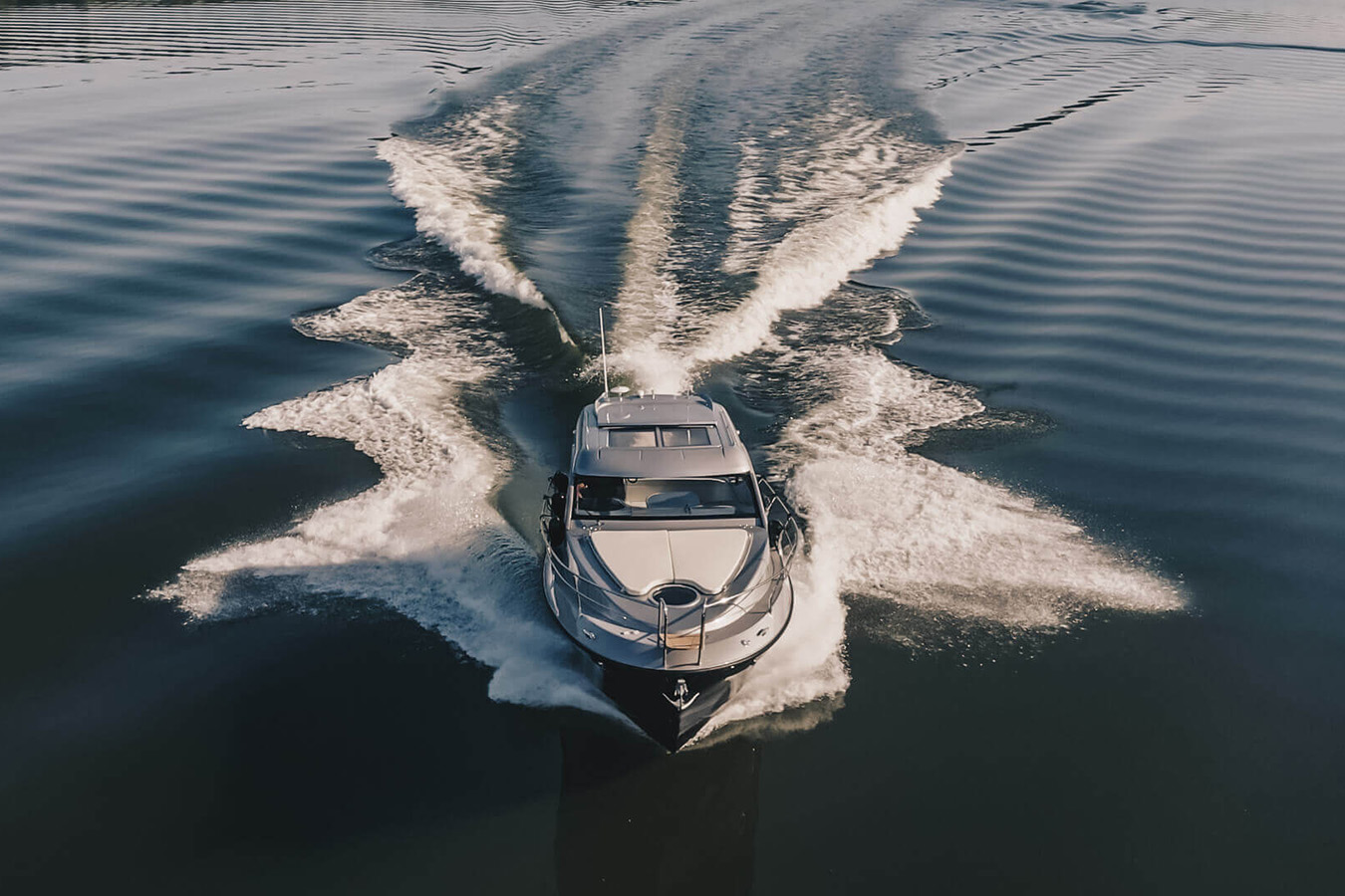 Pogled iz zraka na elegantnu, srebrno-bijelu sportsku jahtu koja brzo plovi mirnom vodom, ostavljajući za sobom širok, pjenast trag