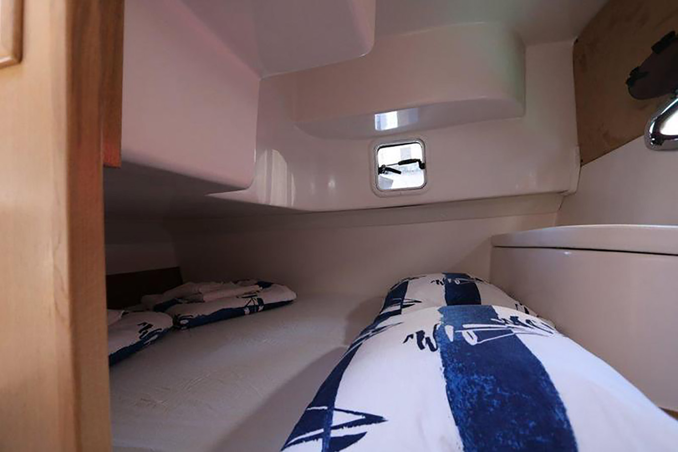 Unutrašnjost kabine sportske jahte, prikazuje dva ležaja s plavo-bijelom posteljinom i malim prozorom