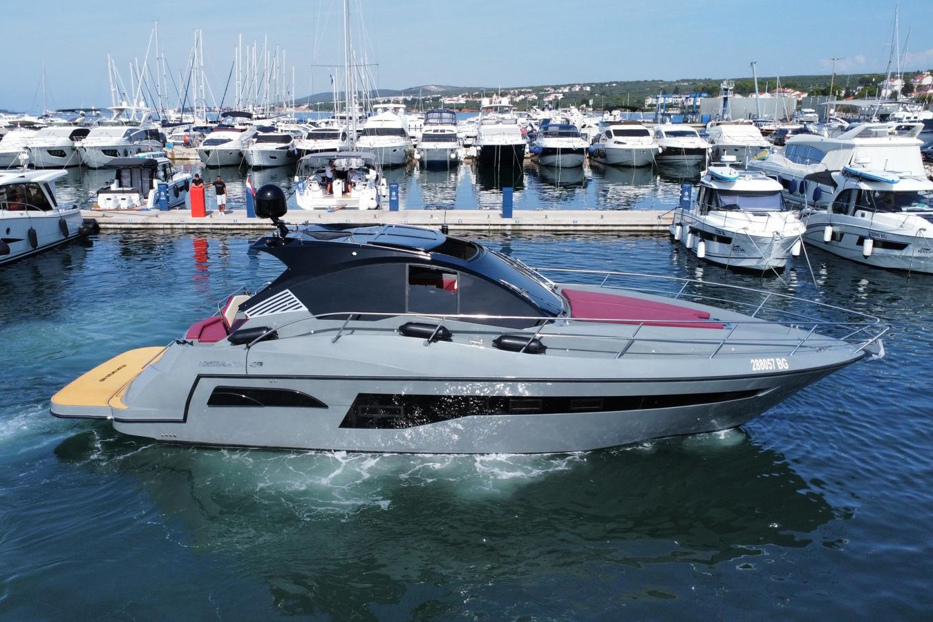 luksuzna jahta u pokretu kroz marinu; sivi trup s crnim hardtopom, crveni jastuci na pramcu i rukohvati od nehrđajućeg čelika, u pozadini niz privezanih jahti i jarbola