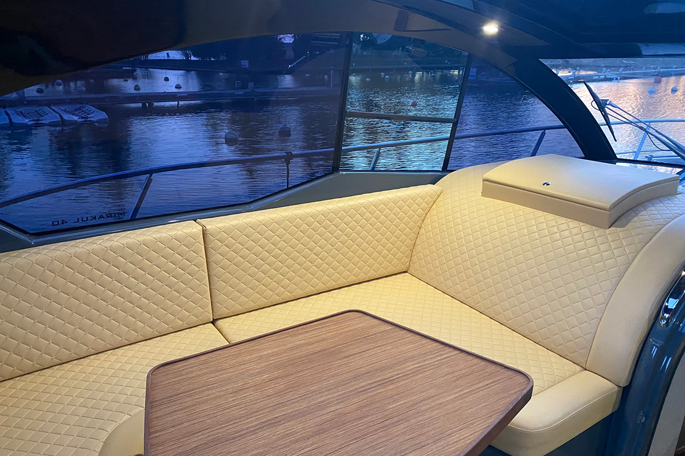 Kokpit luksuzne jahte Mirakul 40 u sumrak: L-sjedeća garnitura presvučena prošivenom bež kožom oko drvenog stola, široka staklena stijenka s pogledom na vez