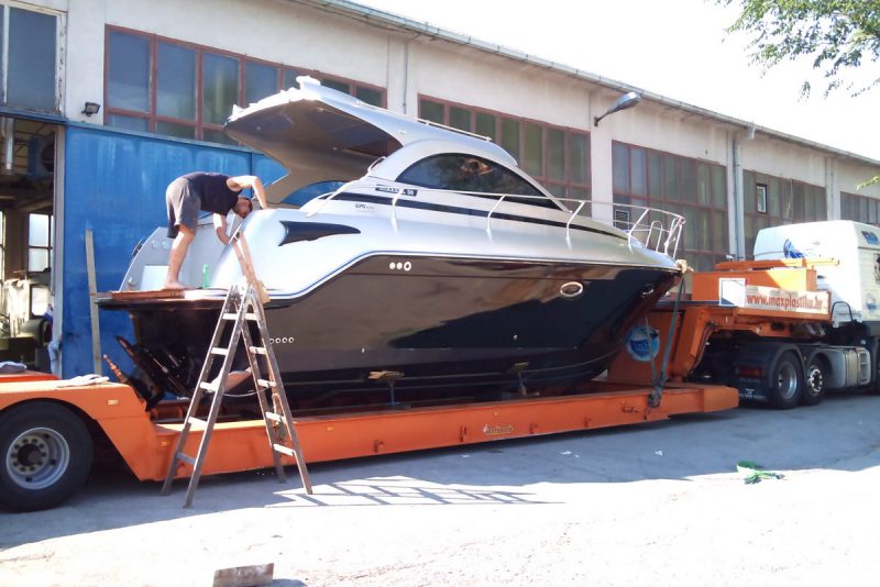 Završna obrada trupa motorne jahte u brodogradilištu; plovilo je na niskopodnoj platformi kamiona spremno za isporuku — realna scena procesa proizvodnje brodova