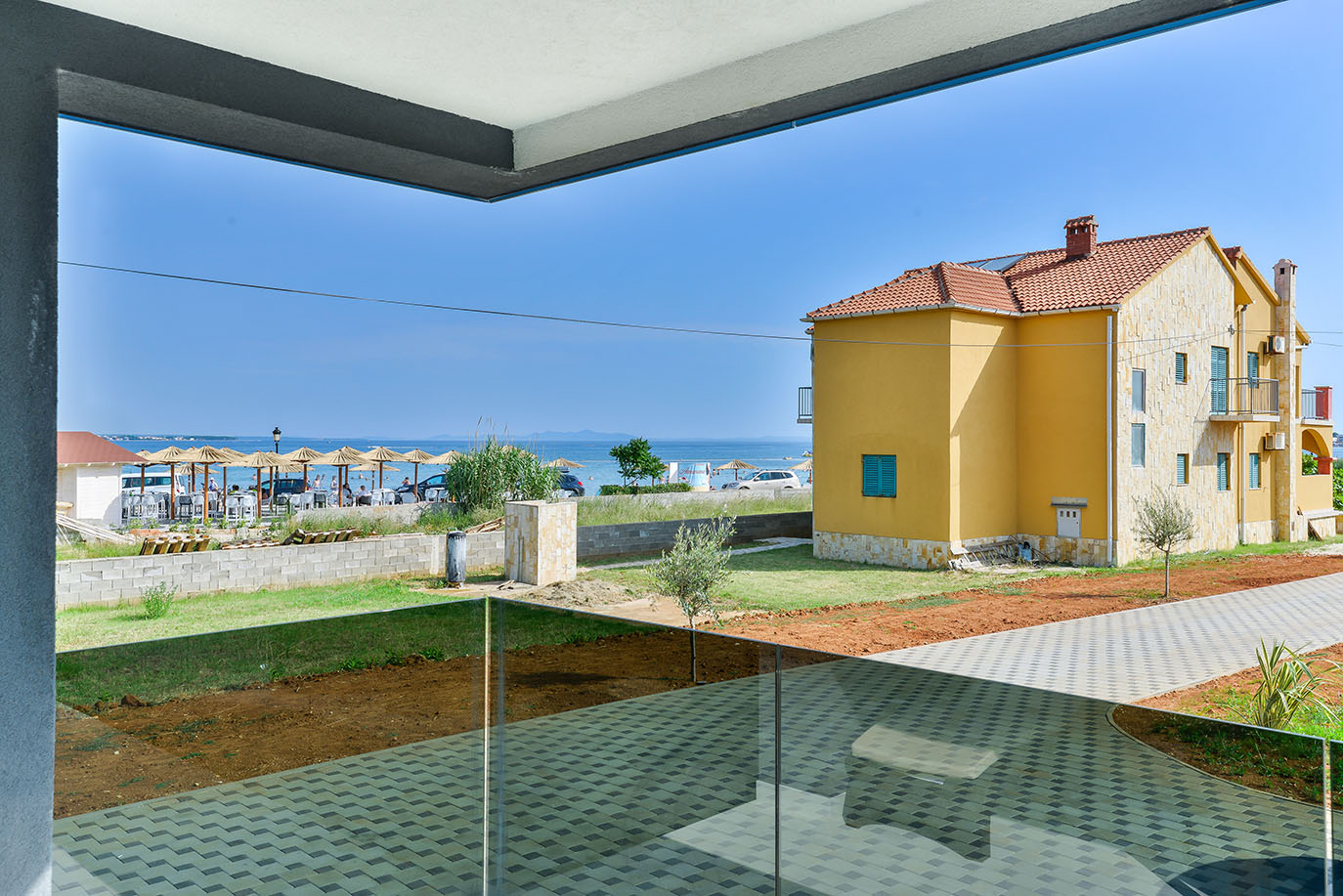Svijetao pogled s modernog balkona na živopisnu žutu kuću s crvenim crijepom, zeleni travnjak i pješčanu plažu sa suncobranima, automobilima i blistavim plavim Jadranskim morem s udaljenim otocima, pružajući primamljiv uvid u apartmane Vir