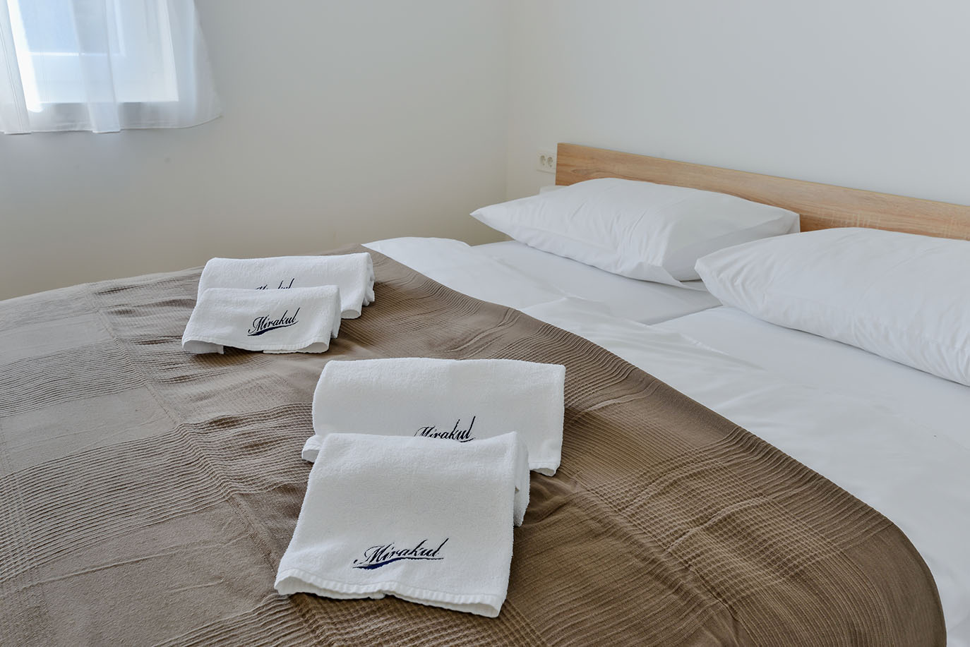 Uredno namješten krevet s bijelim jastucima, smeđom teksturiranom dekom i bijelim ručnicima izvezenim natpisom 'Mirakul', što upućuje na ugodan boravak u Resortu Mirakul
