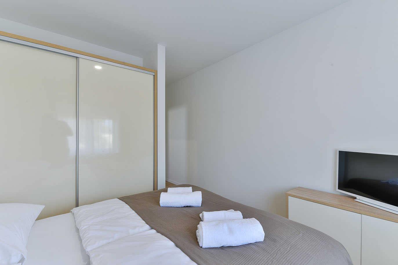 Besprekorna i moderna spavaća soba s udobnim krevetom smeđe-bijele posteljine, uredno složenim bijelim ručnicima, velikim svijetlim kliznim ormarom i televizorom na elegantnom ormariću, što ukazuje na kvalitetan luksuzni smještaj