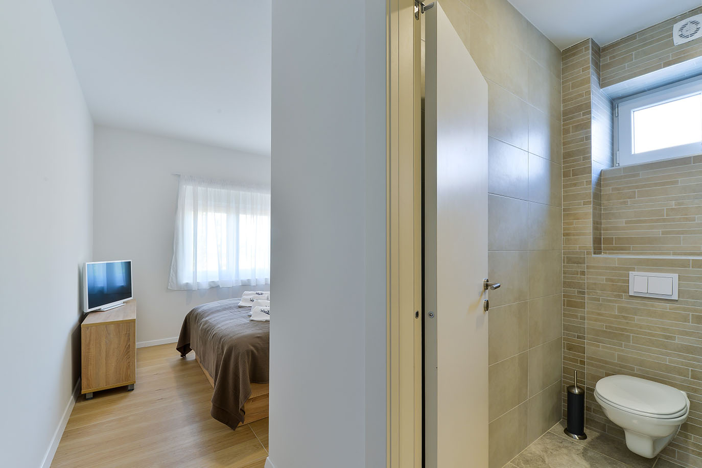 Svijetla i moderna spavaća soba s krevetom i televizorom, koja vodi do elegantne kupaonice s WC-om, nudeći luksuzni smještaj