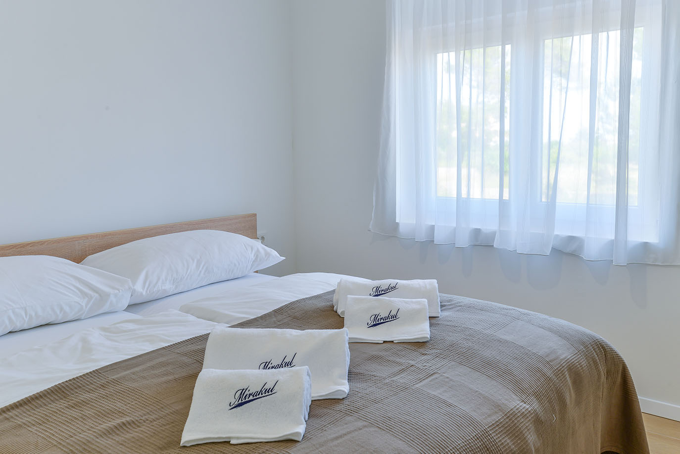 Svijetla spavaća soba u Resortu Mirakul s krevetom, bijelom posteljinom i smeđim prekrivačem, te složenim bijelim ručnicima s izvezenim logom 'Mirakul', pored prozora s prozirnim zavjesama
