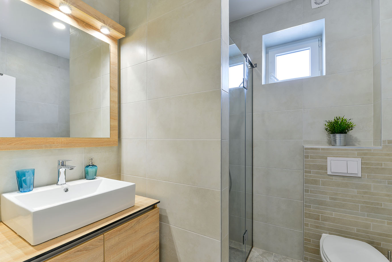 Suvremena kupaonica s elegantnim pravokutnim umivaonikom, velikim ogledalom s drvenim okvirom i rasvjetom, staklenim tušem i svijetlim pločicama, savršena za luksuzni smještaj