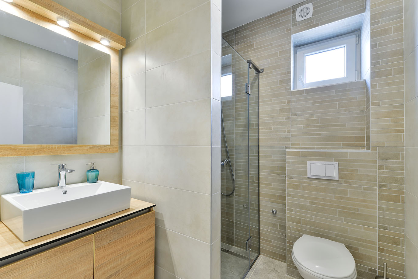 Svijetla, moderna kupaonica s pravokutnim umivaonikom, drvenim ormarićem, osvijetljenim ogledalom, staklenim tušem i zidnim WC-om, idealna za luksuzni smještaj