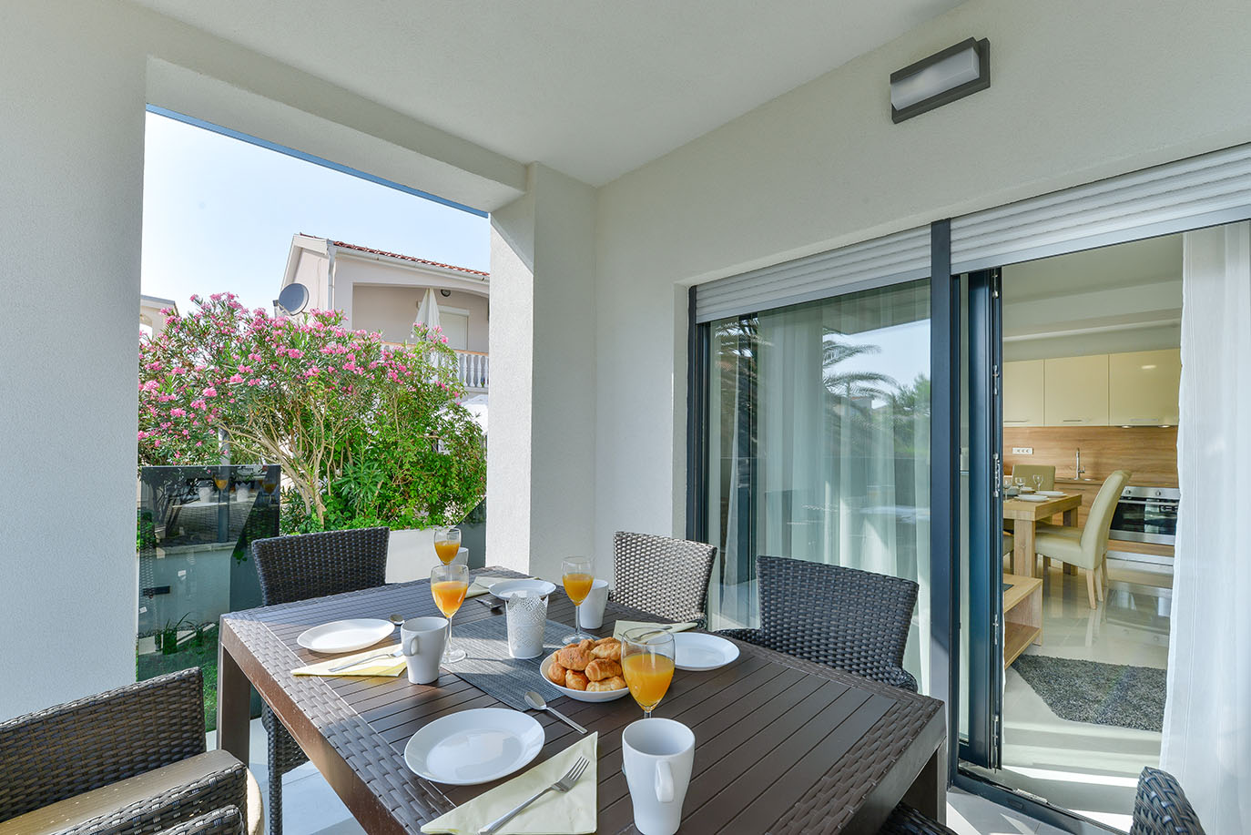 Prekrasno uređena vanjska terasa, dio luksuznog smještaja, s tamnim pletenim stolom za blagovanje i doručkom s narančinim sokom i kroasanima, s pogledom na živopisni vrt