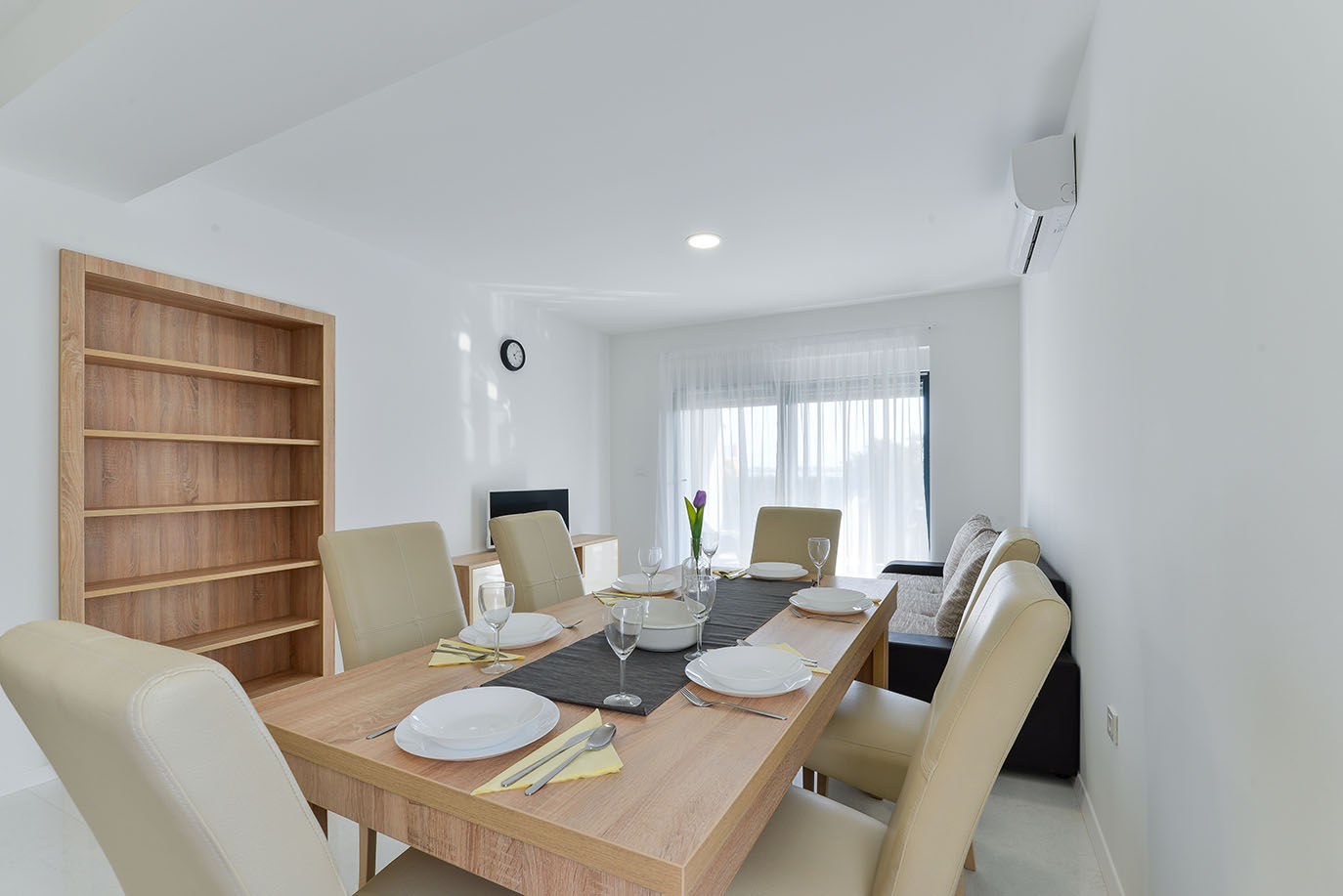 Moderna blagovaonica i dnevni boravak, tipični za apartmane na Viru, s drvenim stolom postavljenim bijelim posuđem, bež stolicama, ugradbenom drvenom policom i udobnom sofom