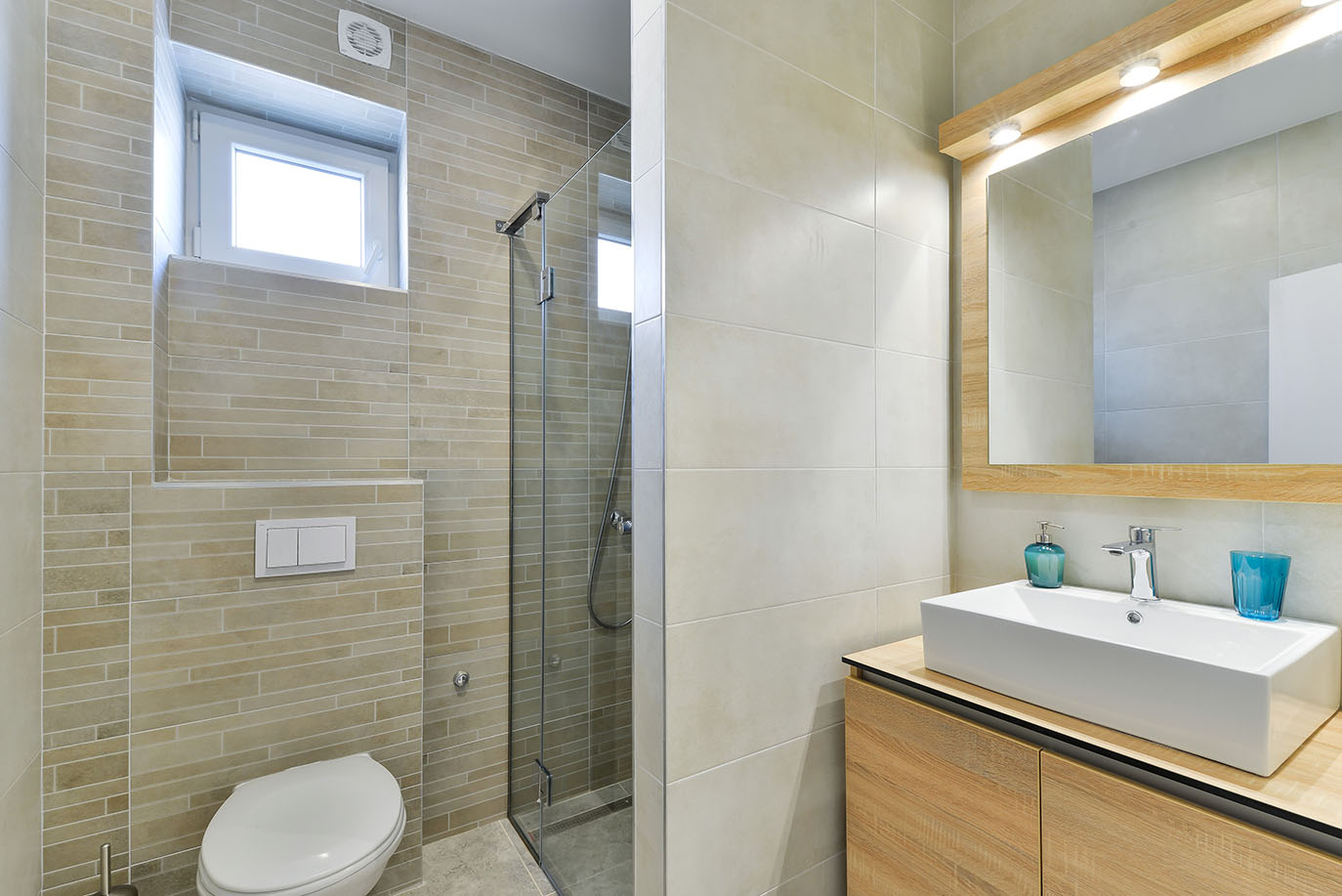 Suvremena kupaonica sa svijetlim pločicama, staklenom tuš kabinom, ormarićem s bijelim umivaonikom i osvijetljenim ogledalom, tipična za luksuzni smještaj