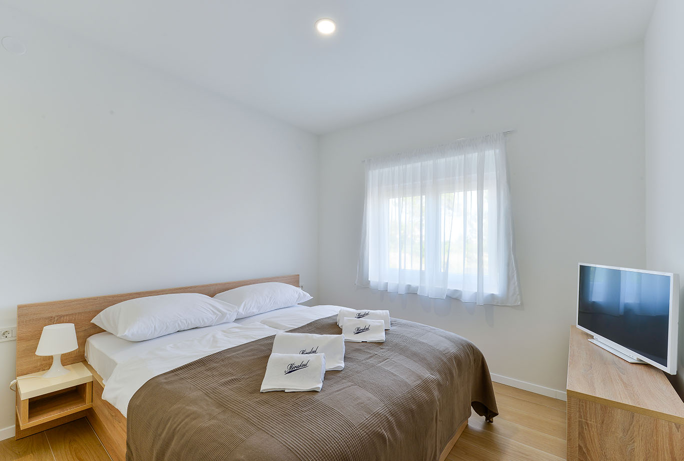 Čista, moderna spavaća soba u Resortu Mirakul s bračnim krevetom, bijelom posteljinom, smeđim pokrivačem i bijelim ručnicima, osvijetljena prirodnim svjetlom s velikog prozora s prozirnim zavjesama