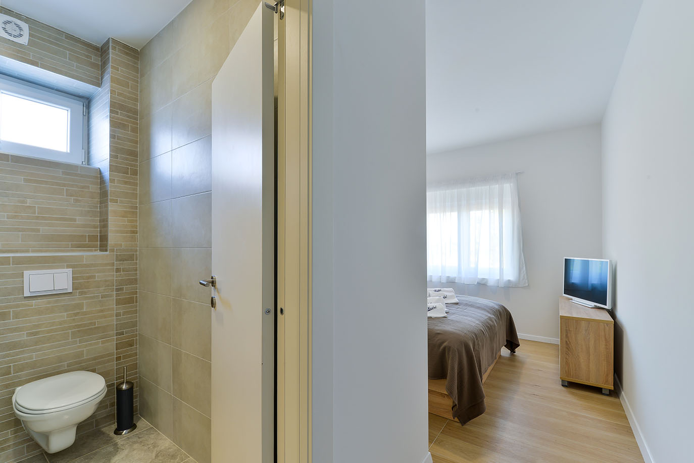 Moderna spavaća soba s krevetom i televizorom pored popločane kupaonice s WC-om, predstavljajući apartmane Vir