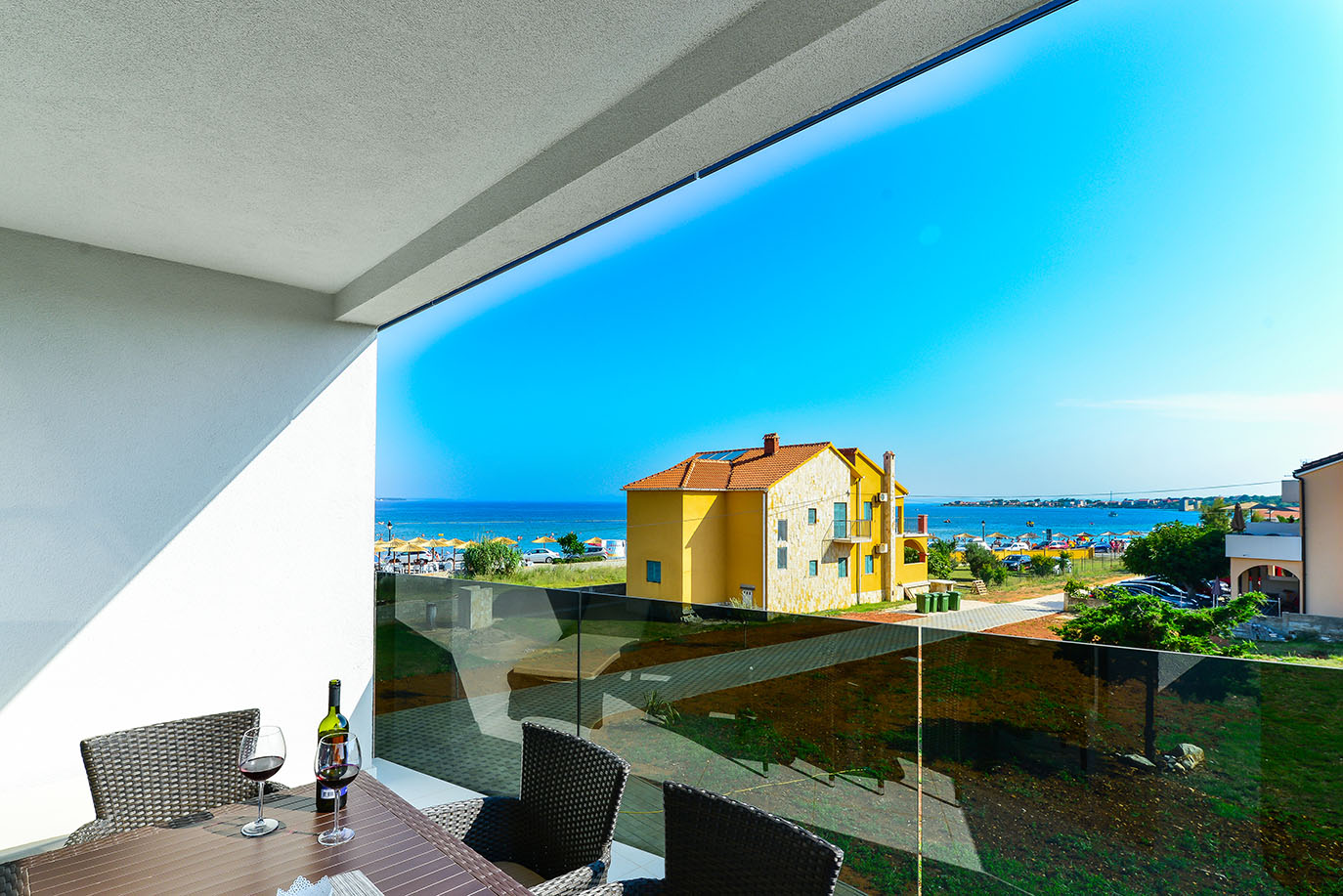 Predivan pogled s balkona na Jadransko more i plažu, snimljen iz apartmana Resorta Mirakul na otoku Viru, sa žutom kućom i suncobranima, te stolom s vinom