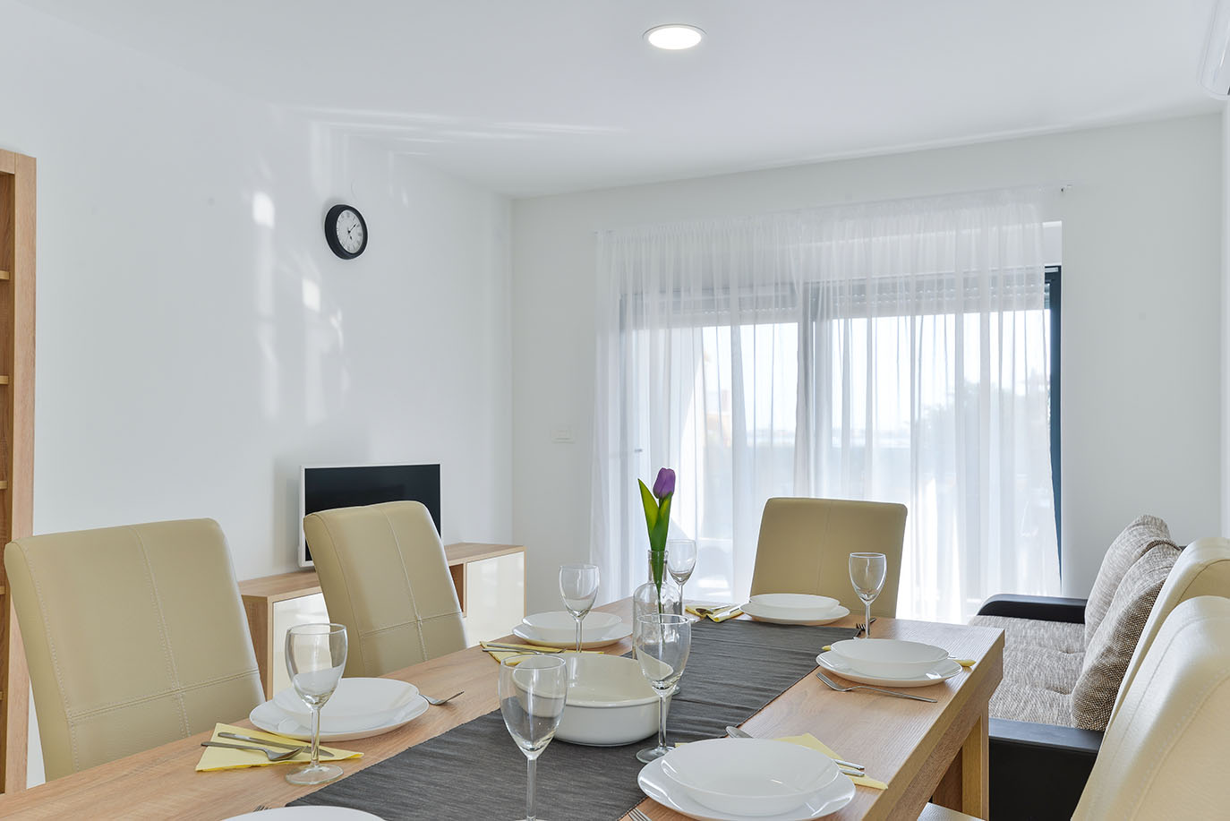Svijetla i moderna blagovaonica u luksuznom smještaju, s drvenim stolom postavljenim za nekoliko gostiju s bijelim tanjurima, čašama i cvjetnim ukrasom, te udobnim bež stolicama i prozorom s prozirnim zavjesama