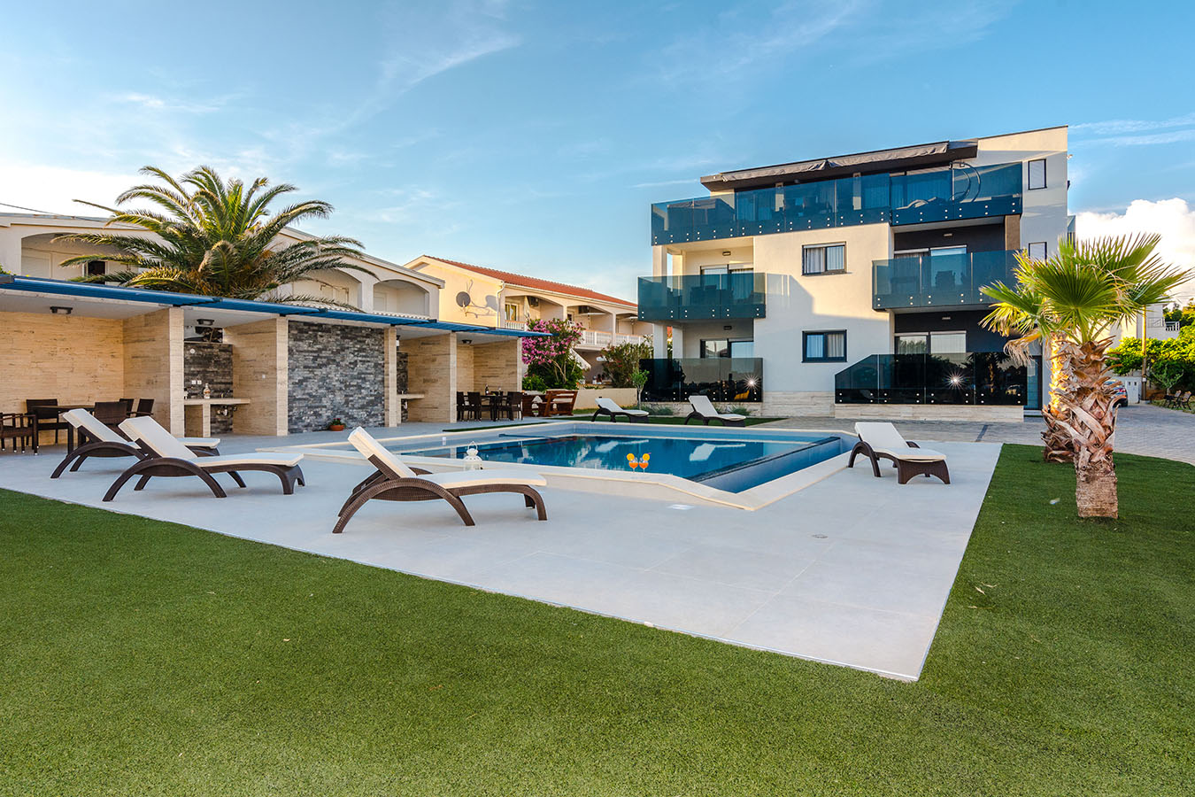 Moderni apartmani Vir s prostranim vanjskim prostorom koji uključuje osvježavajući bazen, udobne ležaljke, natkriveni prostor za objedovanje i bujne palme pod sunčanim nebom