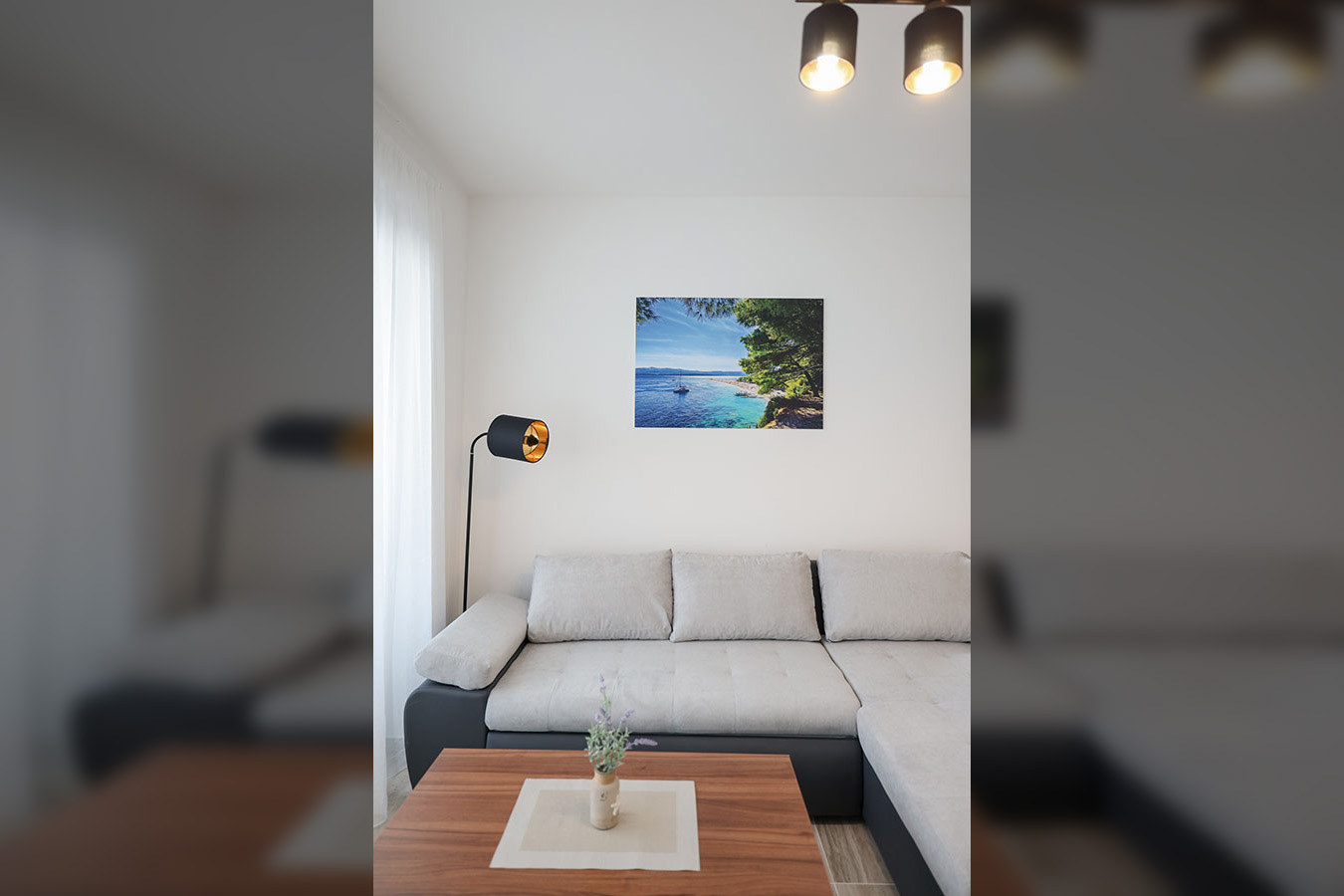 Moderna dnevna soba s udobnom sivom kutnom garniturom, drvenim stolićem i slikom plaže na zidu, idealna za luksuzni smještaj