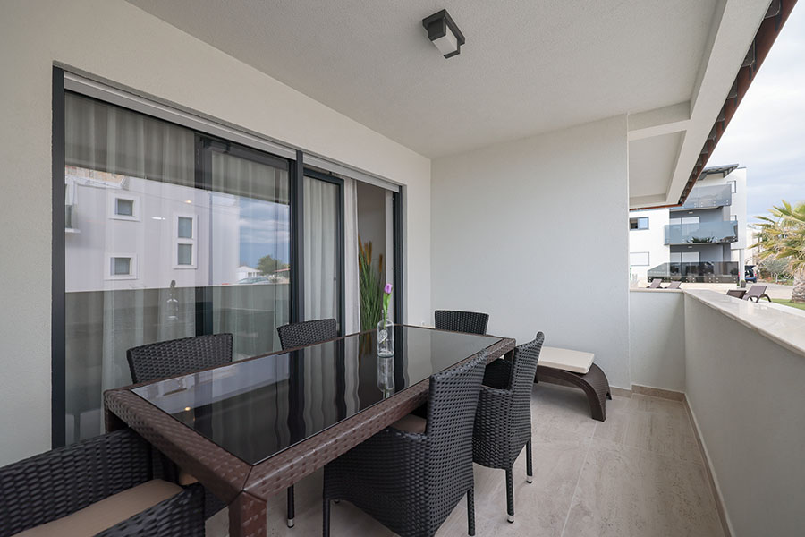 Prostran balkon s modernim vanjskim namještajem, uključujući veliki stol za blagovanje sa staklenom pločom i nekoliko stolica od ratana, pružajući gostima udoban prostor kao dio njihovog luksuznog smještaja