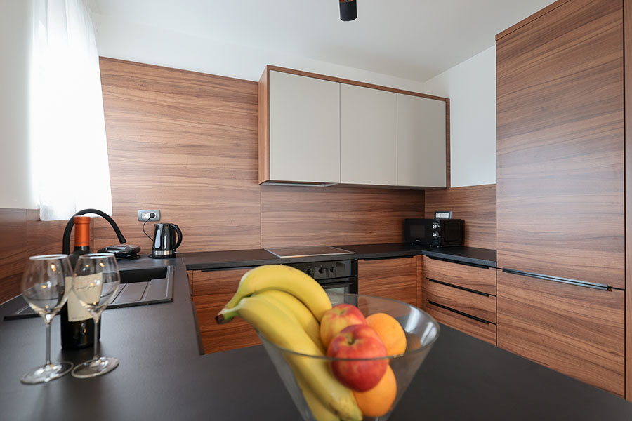 Moderna, dobro opremljena kuhinja s toplim drvenim ormarićima, tamnim radnim pločama, sudoperom, mikrovalnom pećnicom, čašama za vino i zdjelom svježeg voća, naglašavajući kvalitetu luksuznog smještaja