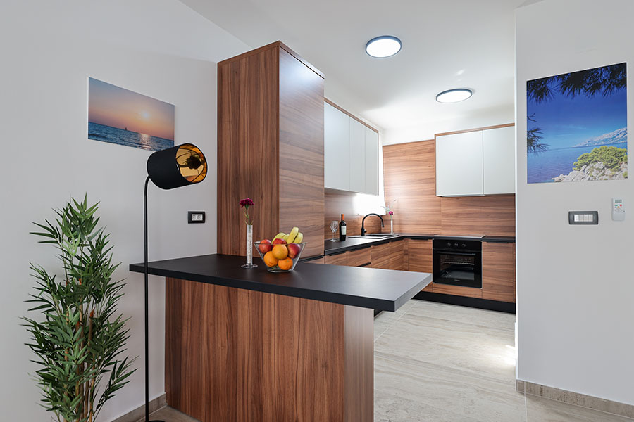 Moderna kuhinja s toplim drvenim ormarićima, bijelim gornjim elementima, crnim radnim pločama i šankom s posudom za voće, prikazujući dizajn luksuznog smještaja
