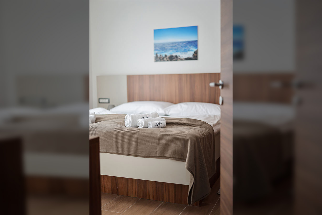 Moderna spavaća soba s udobnim krevetom, bijelom posteljinom i smeđim prekrivačem, idealna za ugodan boravak u apartmanima Vir. Na krevetu su uredno složeni bijeli ručnici, a na zidu iznad uzglavlja visi slika mora