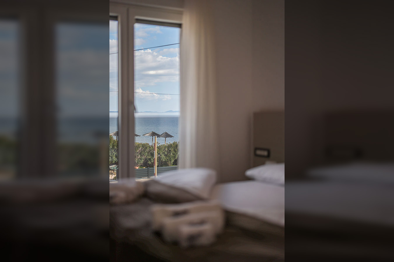 Prekrasan pogled na more s prozora, prikazujući mirno plavo more i daleku obalu, što sugerira luksuzni smještaj s slikovitim pogledom, uključujući dva suncobrana i bujno zelenilo u podnožju