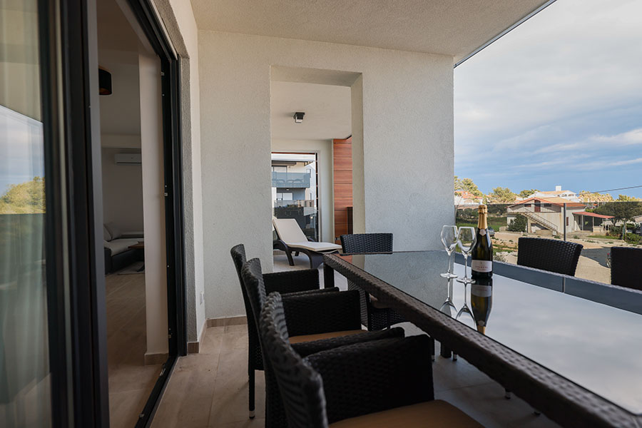 Prostrani moderni balkon sa staklenim blagovaonskim stolom, šampanjcem i čašama, pletenim stolicama i ležaljkom, nudeći savršeno vanjsko okruženje za luksuzni smještaj