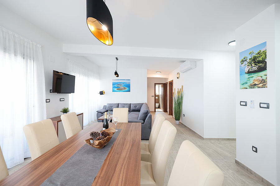 Svijetao i prostran otvoreni dnevni boravak i blagovaonica s modernim drvenim stolom, bež stolicama, sivom kutnom sofom, umjetničkim slikama i zidnim TV-om, prikazujući luksuzni smještaj