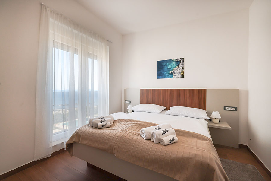 Svijetla spavaća soba u Resortu Mirakul II s velikim krevetom, bijelom posteljinom, bež dekom, smotanim ručnicima i prozorom s prozirnim zavjesama te pogledom na more