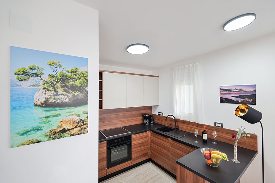 Moderna i potpuno opremljena kuhinja s drvenim donjim elementima, bijelim gornjim elementima, crnom radnom pločom i šankom s voćem, idealna za kvalitetne apartmane na Viru