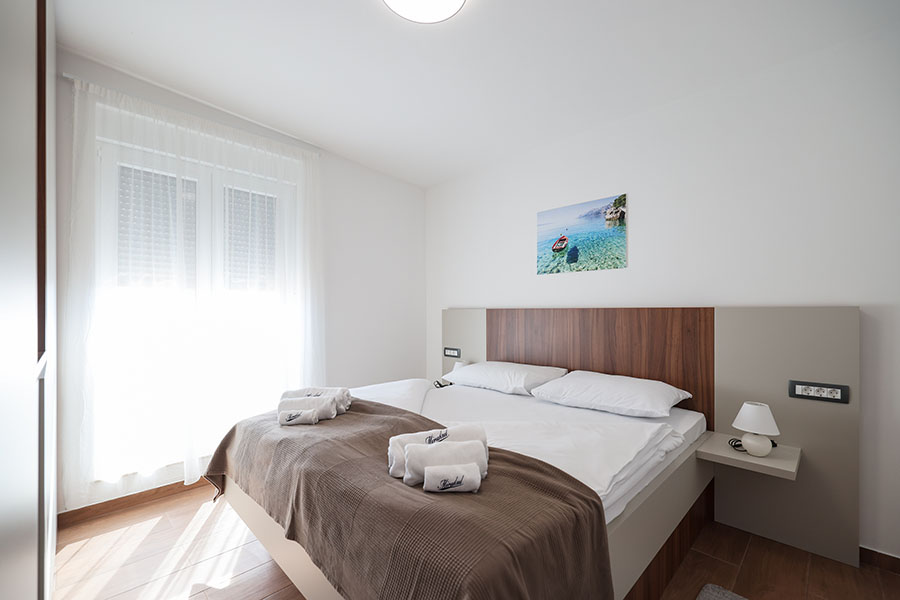 Svijetla i moderna spavaća soba s velikim krevetom, bijelom posteljinom, smeđim prekrivačem, smotanim bijelim ručnicima, noćnim ormarićem s lampom i prozorom s prozirnim zavjesama, idealna za luksuzni smještaj