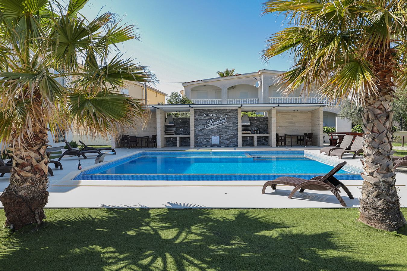 Sunčano vanjsko područje bazena u Resortu Mirakul, s udobnim smeđim ležaljkama, bujnom zelenom travom i visokim palmama pod vedrim plavim nebom, s modernom zgradom i roštiljem u pozadini