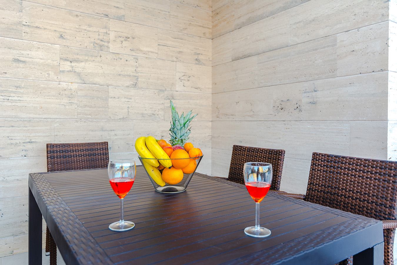 Vanjski prostor za sjedenje s modernim stolom, zdjelom punom voća i dva pića, te elegantnim kamenim zidom u pozadini, predstavljajući luksuzni smještaj
