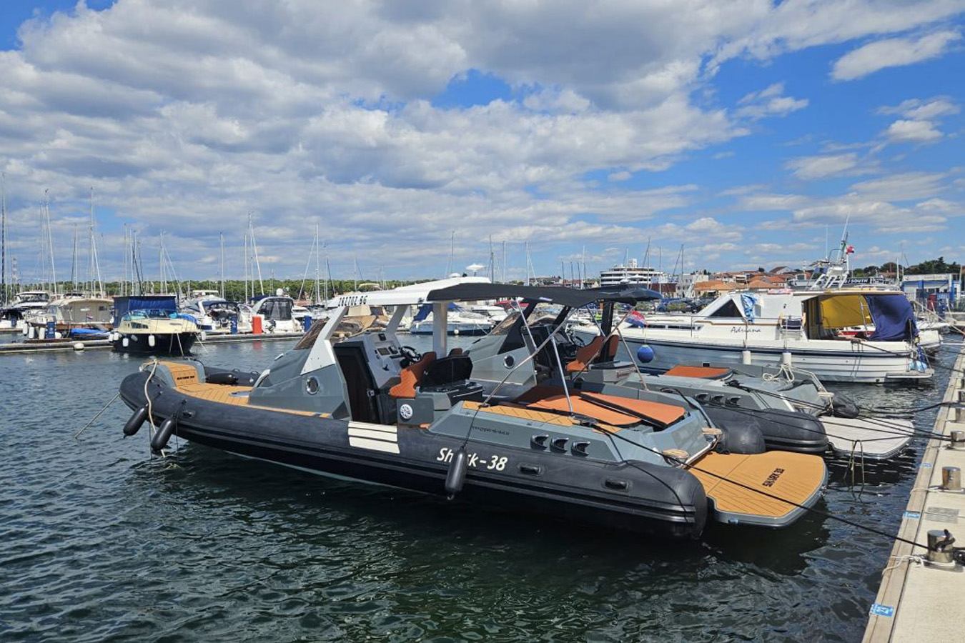 Shark 38 RIB privezan u marini, tamnosivi tubusi, narančasti jastuci, T-top nadgrađe i tikove platforme; robustan, premium dizajn za brza transfer plovila