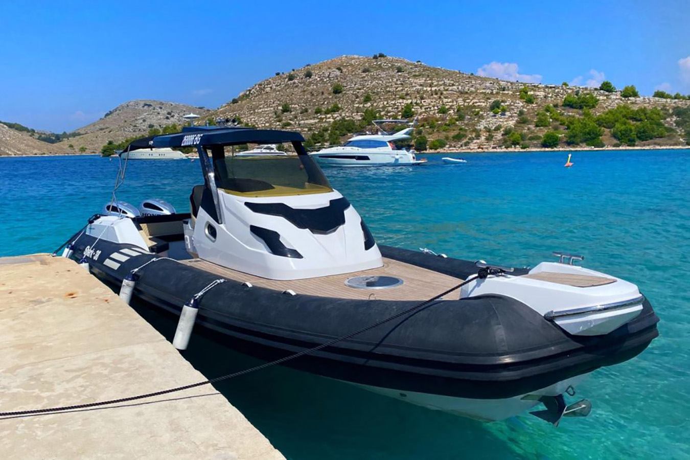 Elegantan crno-bijeli luksuzni gumenjak usidren je uz betonski pristan u uvali s kristalno čistim tirkiznim morem, stjenovitim otocima i jahtom u pozadini pod vedrim plavim nebom