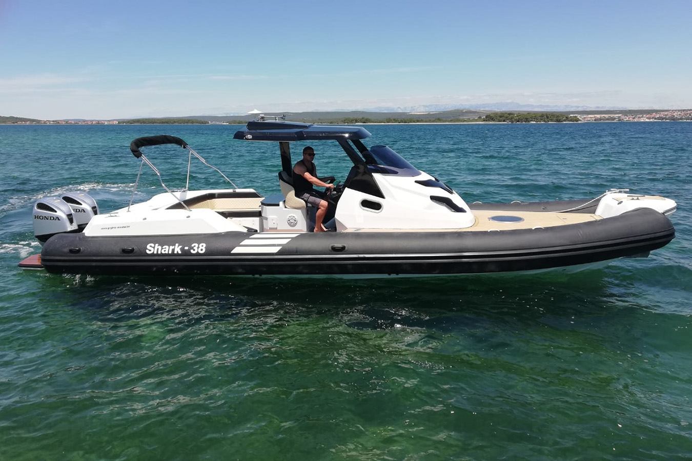 Muškarac upravlja modernim crno-bijelim brodom Shark 38 s dva Honda motora na bistrom plavom moru, s obalom otoka u pozadini pod sunčanim nebom