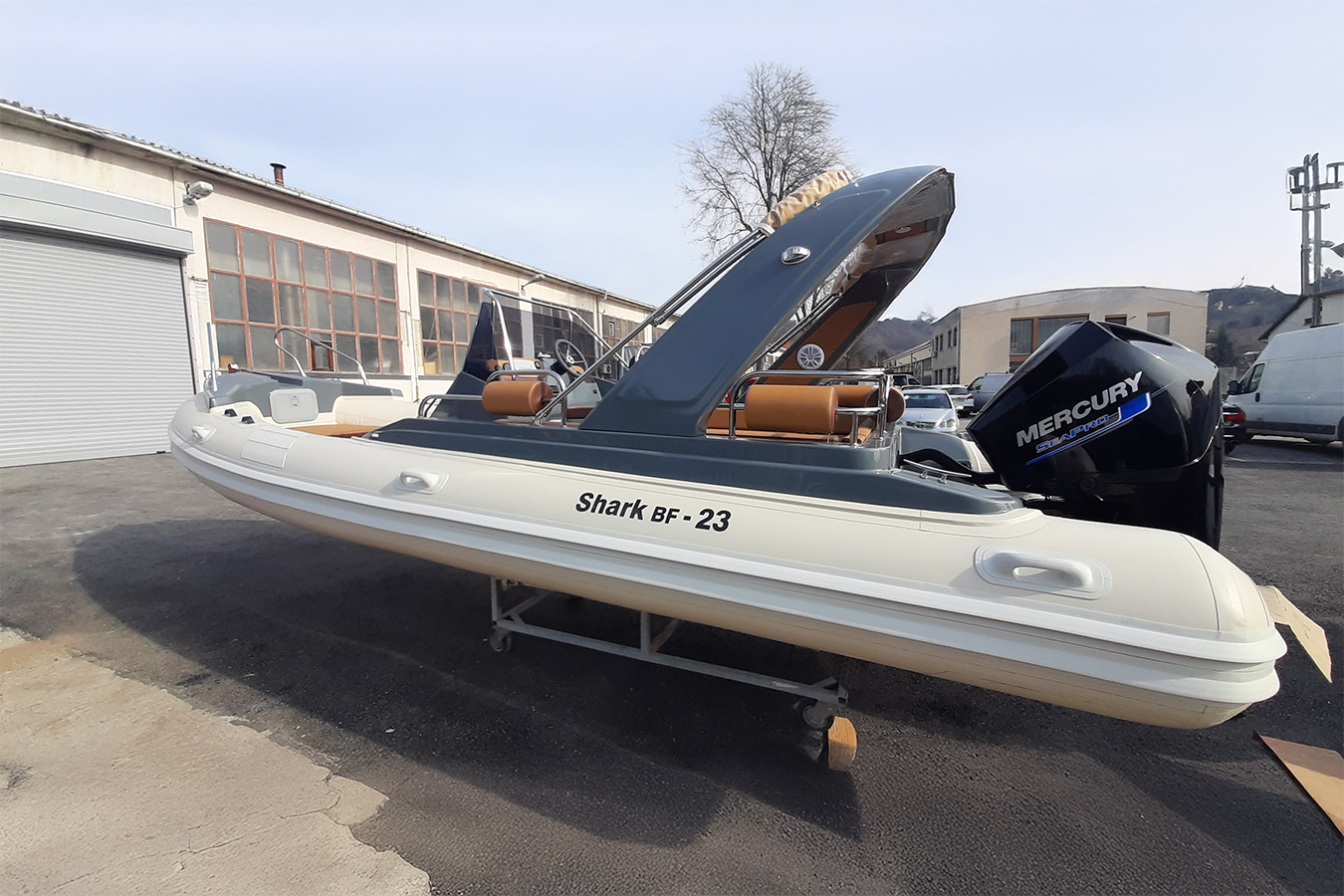 Luksuzni gumenjak Shark BF-23 bijele boje, s tamno sivom tendom i smeđim sjedalima, stoji na postolju na otvorenom s vanbrodskim motorom Mercury Seapro