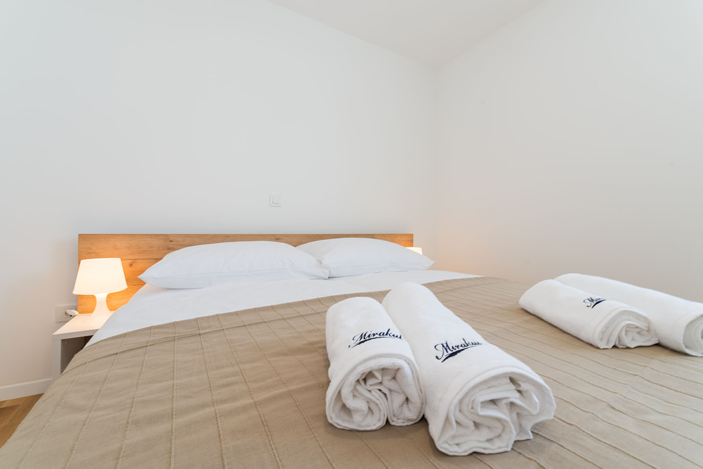 Uredna i udobna spavaća soba u apartmanima Villa Mirakul, s udobnim bračnim krevetom, prekrivačem neutralne boje, noćnom lampom i svježe smotanim ručnicima