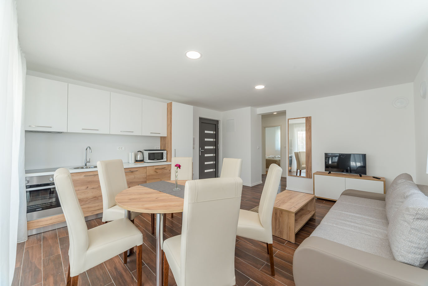 vijetli i moderni otvoreni prostor kuhinje, blagovaonice i dnevnog boravka, koji prikazuje udoban prostor idealan za smještaj u apartmani Vir, s potpuno opremljenom kuhinjom s bijelim ormarićima, okruglim drvenim stolom za ručavanje s krem stolicama i udobnim kaučem s TV prostorom