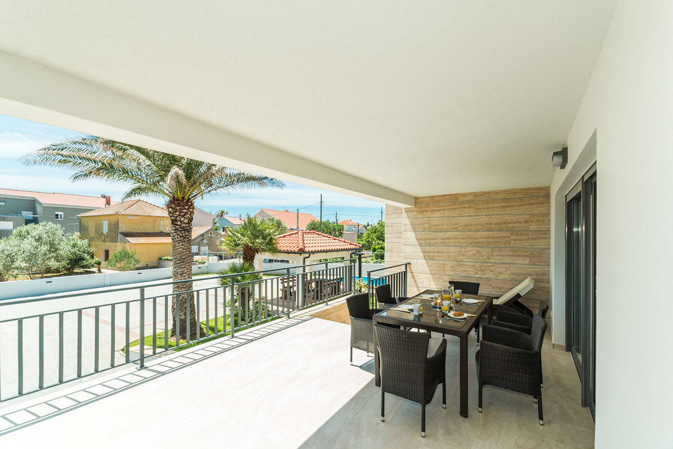 Prostrani natkriveni balkon s postavljenim stolom za doručak i udobnim sjedalima, s pogledom na palme i stambene zgrade, savršen za uživanje u apartmani Vir