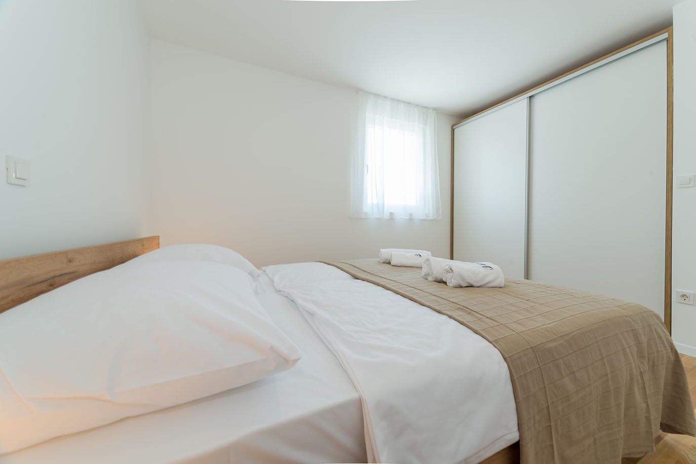 Moderna i uredna spavaća soba s udobnim krevetom, bijelom posteljinom, bež dekom, svježim ručnicima, drvenim uzglavljem i prostranim kliznim ormarom, idealna za apartmane Vir
