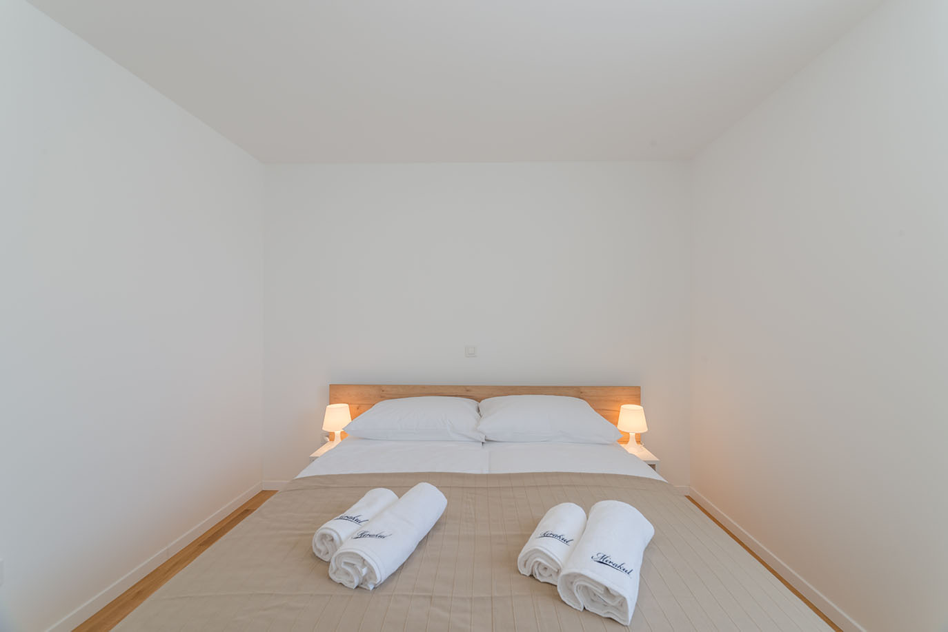 Prostrana i svijetla spavaća soba s bračnim krevetom, bijelom posteljinom, dvjema noćnim lampama i uredno složenim bijelim ručnicima s logom 'Mirakul', odražavajući čistoću i udobnost u Villa Mirakul