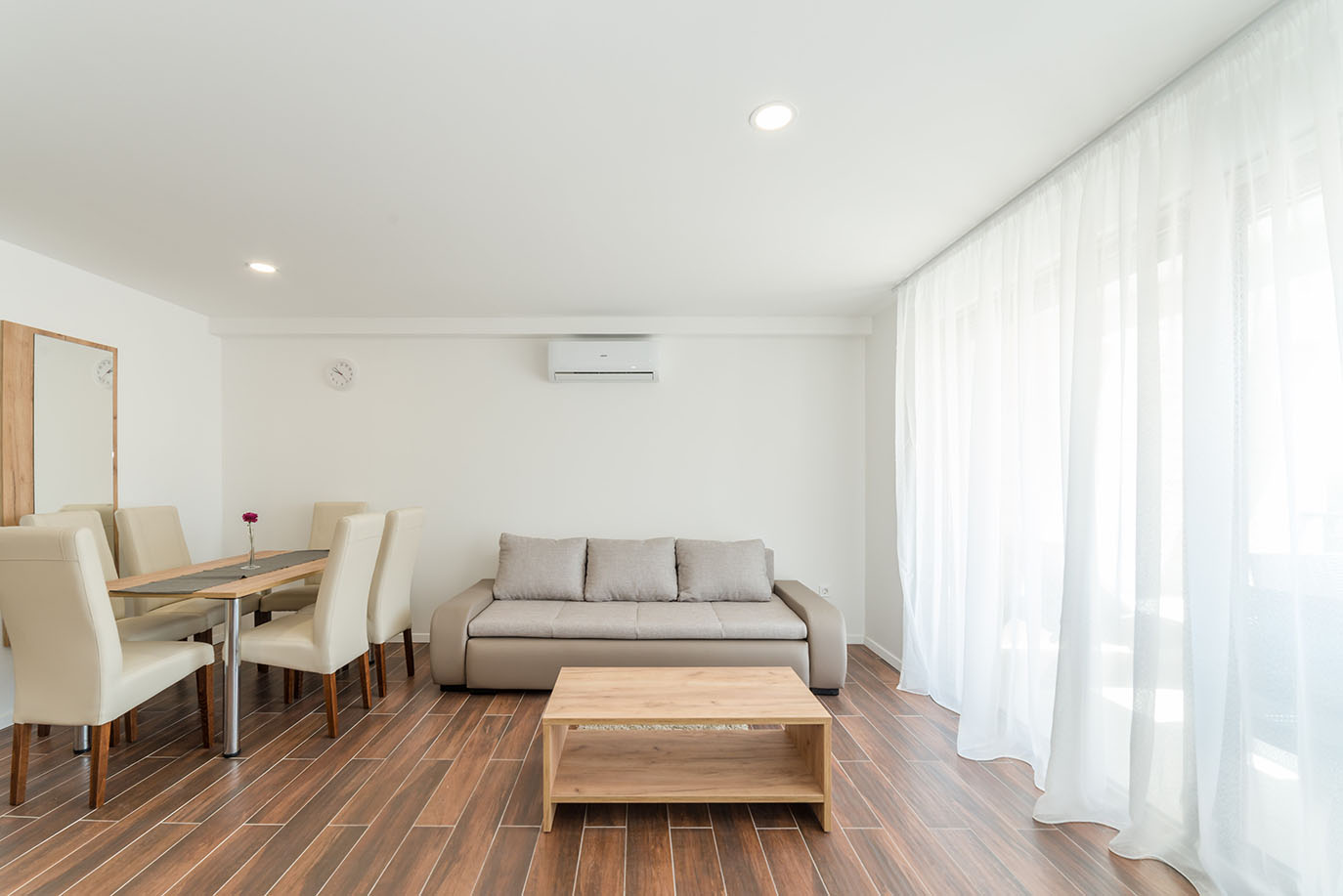 Svijetli i moderni dnevni boravak s blagovaonicom, tipičan za udoban smještaj kao što su apartmani Vir, opremljen udobnom sofom, drvenim stolićem, blagovaonskim stolom sa svijetlim stolicama, drvenim podom i prozorima s prozračnim bijelim zavjesama