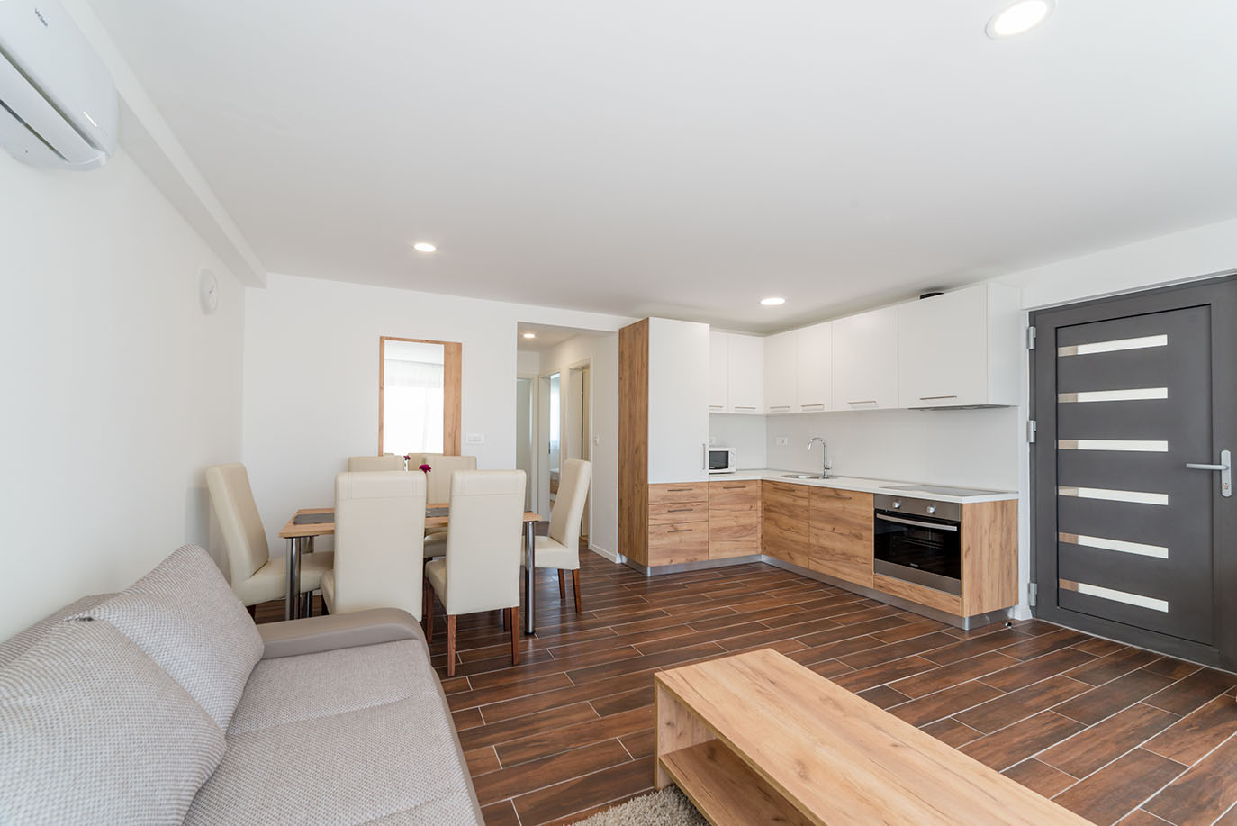 Svijetao, moderan otvoreni dnevni boravak i kuhinja, tipičan za apartmane Vir, s kuhinjom u svijetlom drvu i bijelim elementima, blagovaonskim stolom s bež stolicama i udobnom sivom sofom