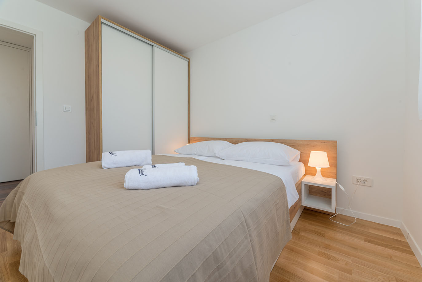 Moderna spavaća soba u Villa Mirakul s velikim krevetom, uzglavljem od svijetlog drveta, čistom bijelom posteljinom i dva smotana ručnika, pored bijelog noćnog ormarića s lampom i prostranog ormara