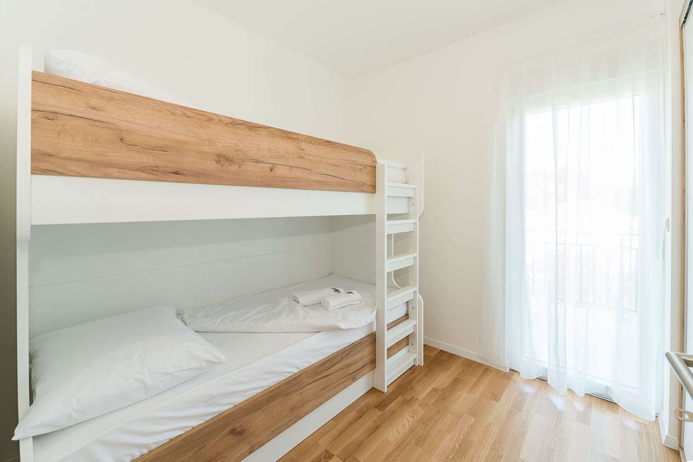 Svijetla spavaća soba s modernim bijelim krevetima na kat i detaljima od prirodnog drva, svježom bijelom posteljinom i velikim prozorom, idealna za obitelji koje borave u apartmani Vir