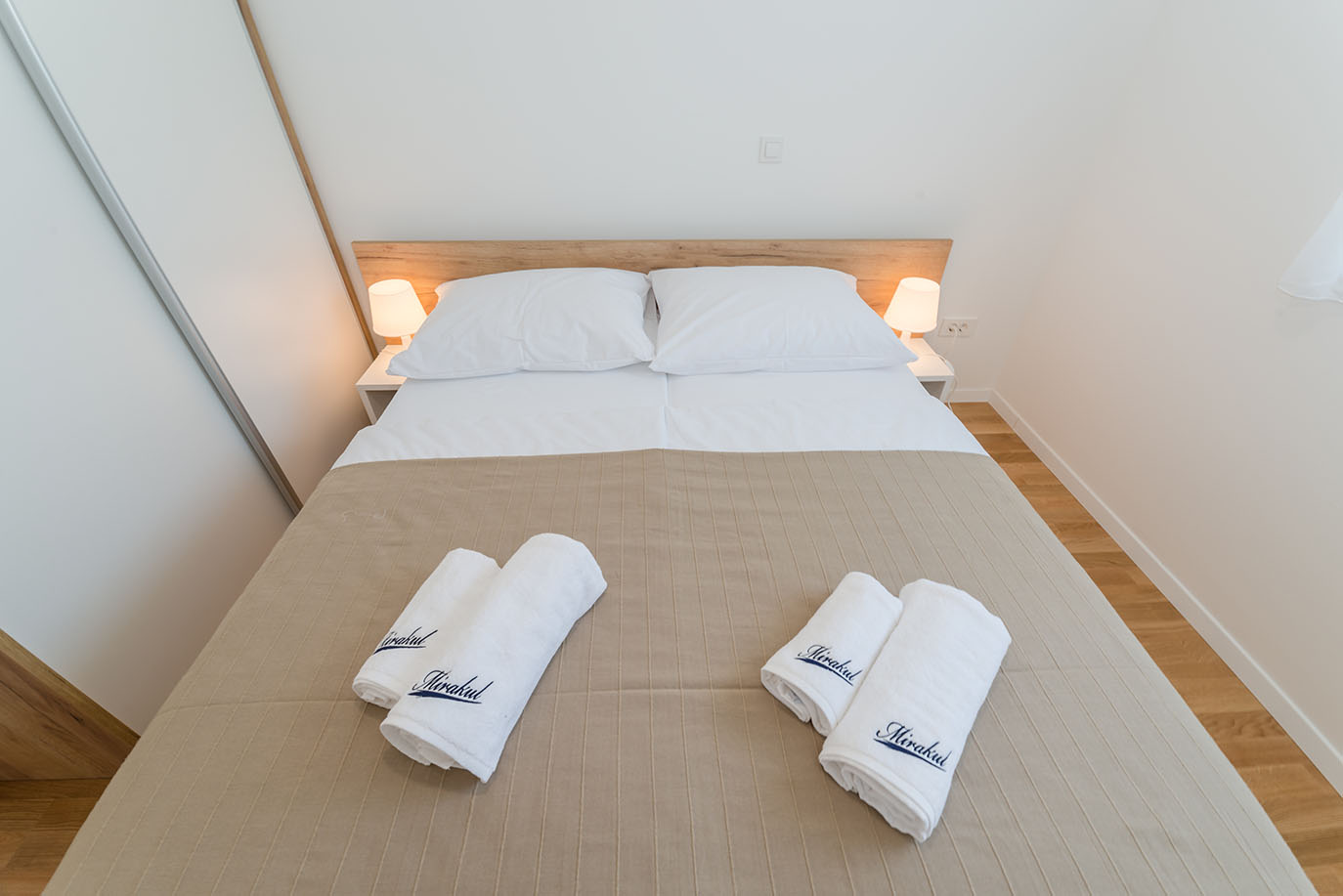 Udobna dvokrevetna spavaća soba u apartmanima Vir, opremljena bijelom posteljinom, bež prekrivačem, noćnim ormarićima s lampama te uredno složenim bijelim ručnicima s natpisom Mirakul