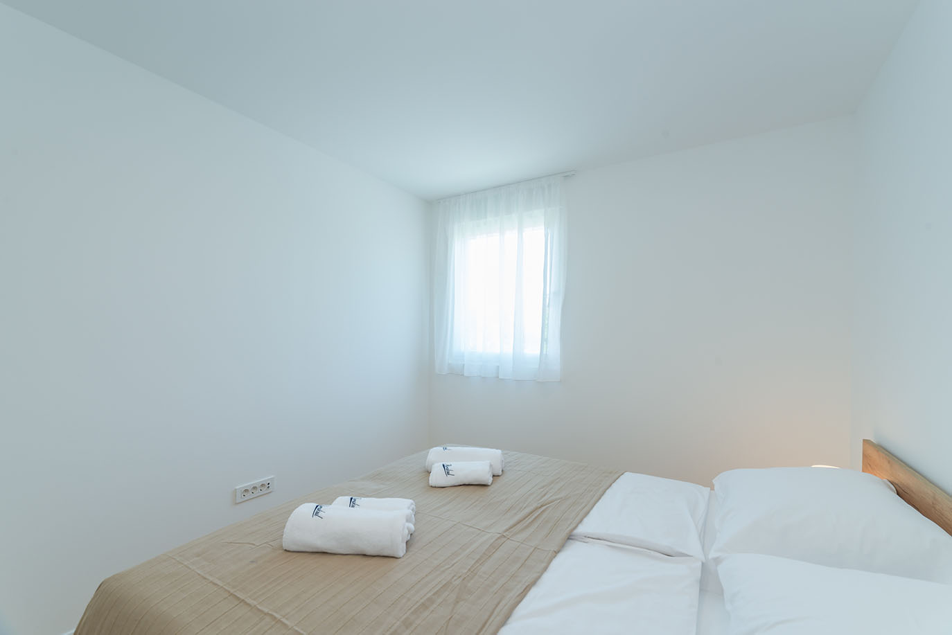 Svijetla i minimalistička spavaća soba s udobnim krevetom, bijelom posteljinom, bež prekrivačem i uredno složenim bijelim ručnicima, tipična za apartmane Vir. Prozor s prozirnim zavjesama propušta obilje prirodnog svjetla