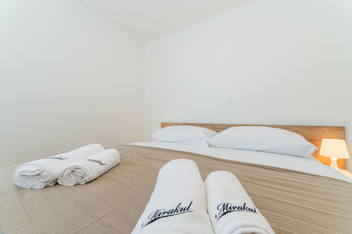 Svijetla spavaća soba u Villa Mirakul, s uredno namještenim krevetom, bijelim jastucima, bež dekom i smotanim bijelim ručnicima s natpisom 'Mirakul', pored noćne lampe