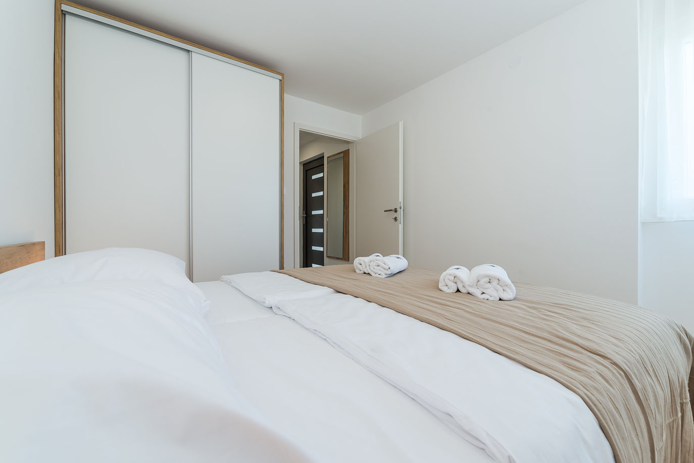 Moderna i uredna spavaća soba s udobnim krevetom, bijelom posteljinom, bež prekrivačem i svježim smotanim ručnicima, tipična za kvalitetne apartmane na Viru